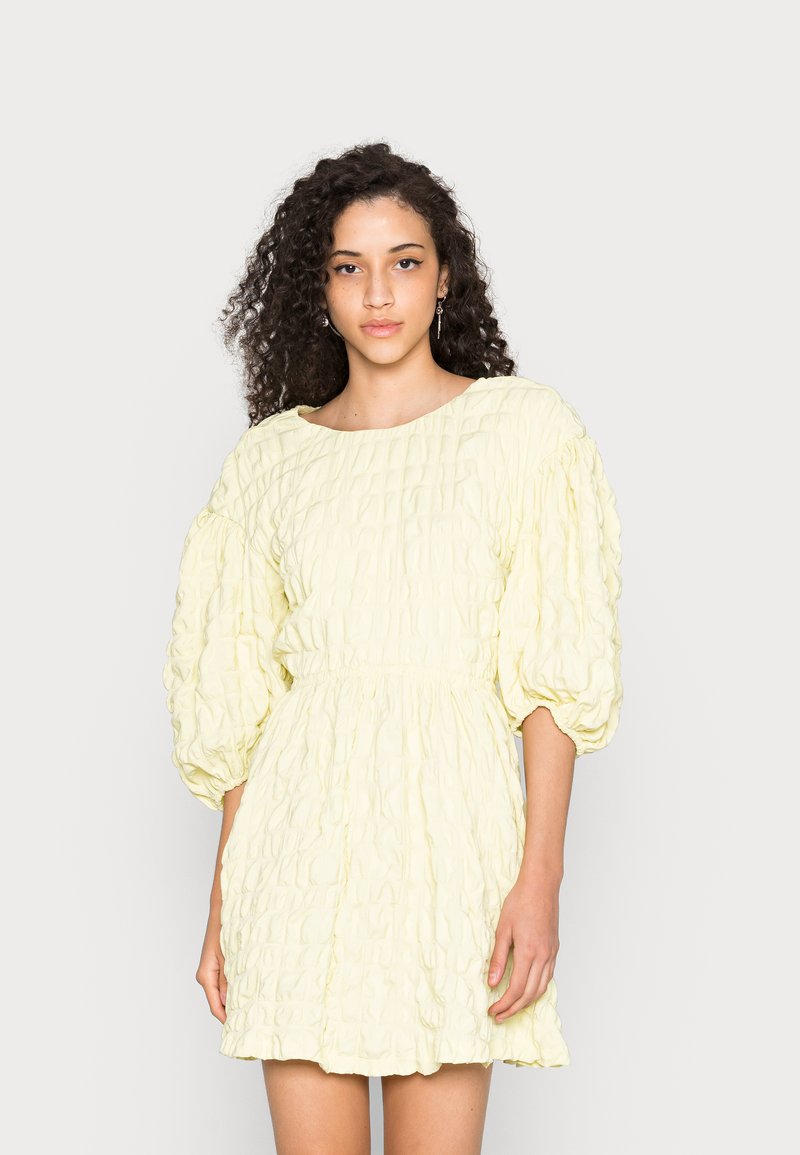 Vestido mini amarelo, texturizado, com cintura franzida, decote redondo e mangas bufantes. Tecido suave com um padrão de pregas subtil.