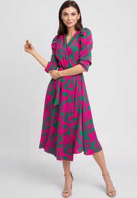 Kwiatowa sukienka typu wrap w jasnej zieleni z dużymi różowymi kwiatowymi wzorami, krótkimi bufkami i paskiem do wiązania; długość do łydki z lekko opływającą sylwetką.