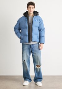Puffer jacket in light blue with black hood, featuring zip closure and two front pockets; worn with distressed light blue jeans and white sneakers.