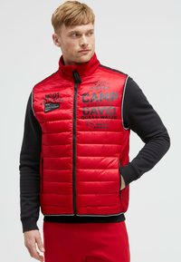 Rote gepolsterte Weste mit schwarzen Akzenten, Reißverschlusstaschen und bedrucktem Text. Sie verfügt über einen hohen Kragen und gerippte Bündchen für zusätzliche Wärme.