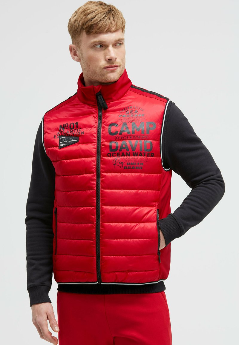 Rote gepolsterte Weste mit schwarzen Akzenten, Reißverschlusstaschen und bedrucktem Text. Sie verfügt über einen hohen Kragen und gerippte Bündchen für zusätzliche Wärme.