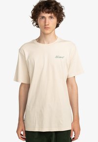Beige cotton T-shirt with short sleeves, featuring a green logo graphic on the left chest. Classic crew neck design.