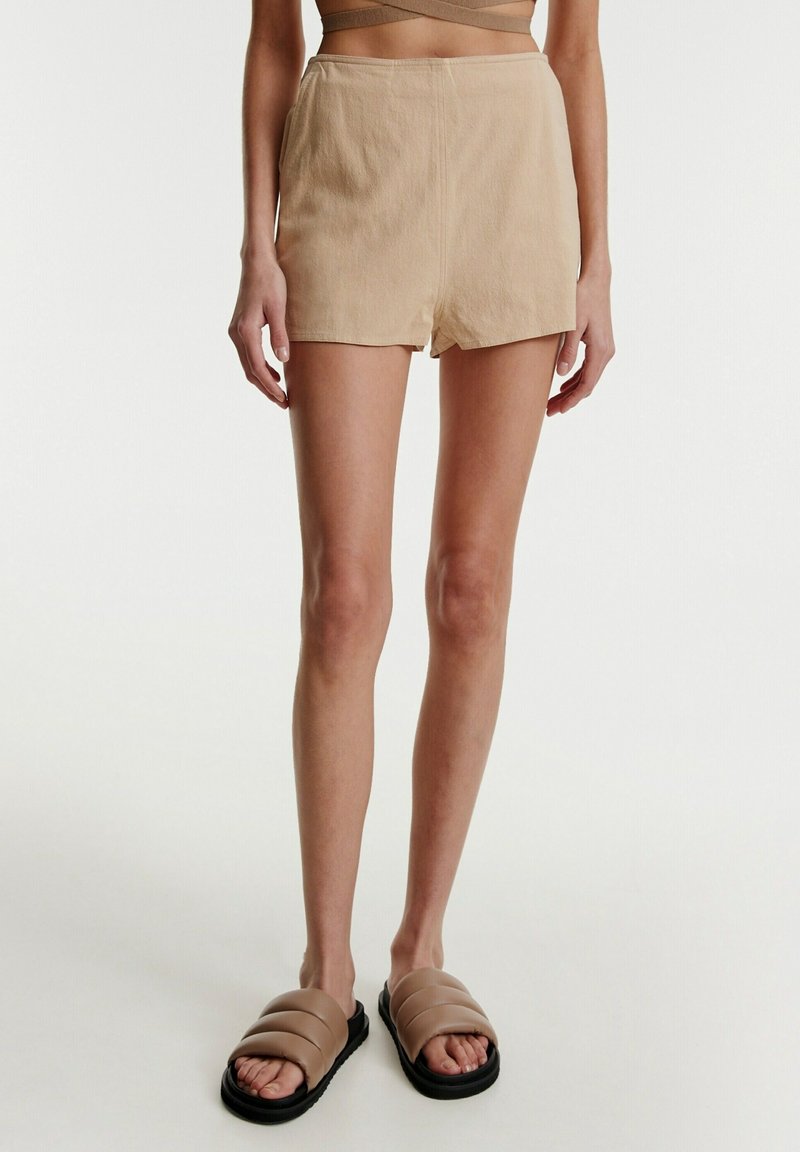 Femme portant un short beige taille haute avec fentes latérales et des sandales slides rembourrées beige, debout devant un fond blanc.