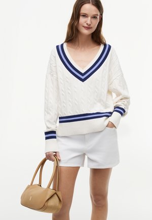 Femme portant un pull blanc en maille torsadée avec des rayures bleues, un short blanc, tenant un sac à main en cuir marron par les poignées.