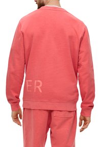 Sweat-shirt corail avec un col rond et des poignets côtelés. Présente un détail embossé "ER" au dos. Texture douce, coupe décontractée.