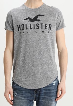 Persoon draagt een grijs Hollister California T-shirt met een zwart meeuwenlogo, gecombineerd met blauwe versleten spijkerbroek.