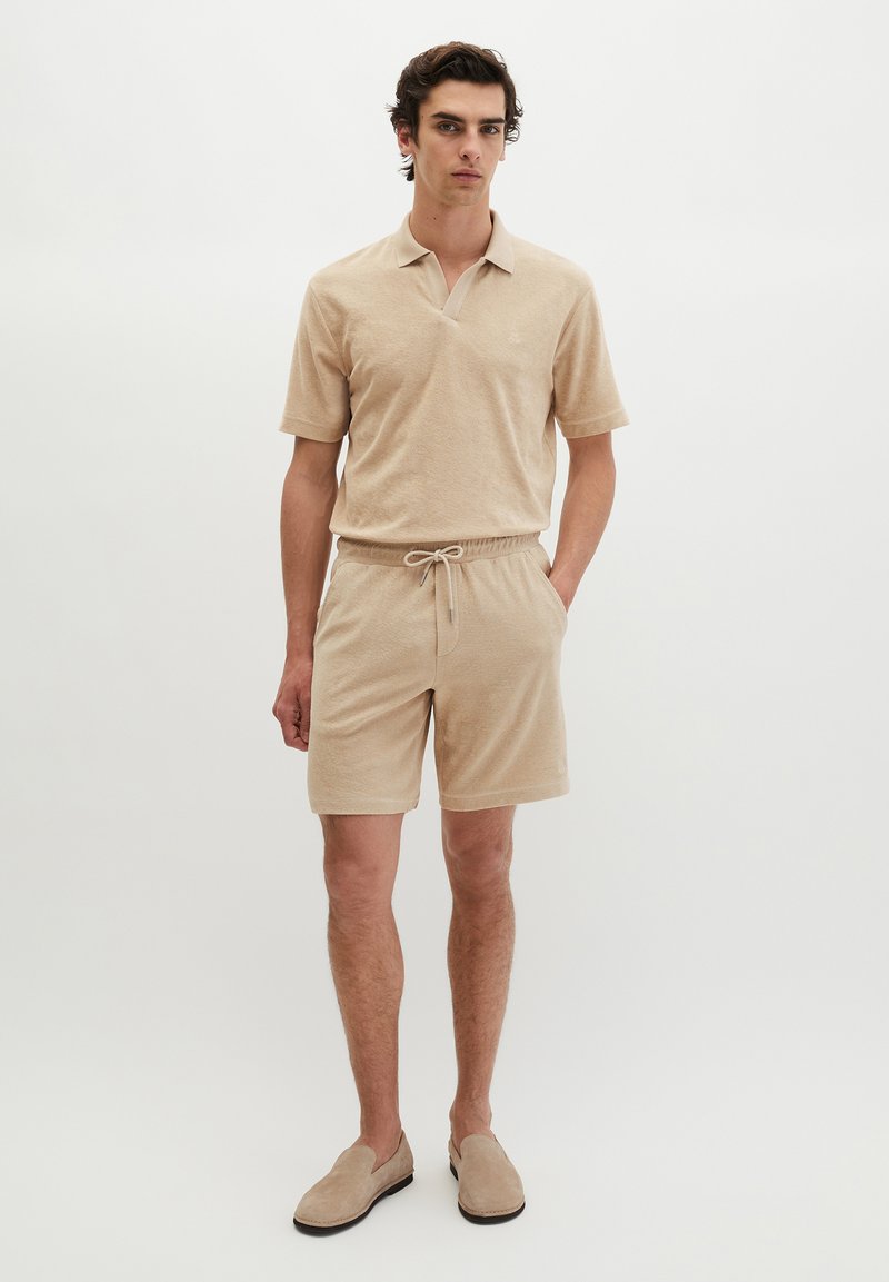 Jeune homme debout, les mains dans les poches, portant un polo beige, un short à cordon assorti et des chaussures slip-on sur fond blanc uni.
