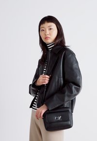 Schwarze Lederjacke mit einem kragenförmigen Design, kombiniert mit einem gestreiften Rollkragenpullover. Eine kleine schwarze Umhängetasche mit einem silbernen Logo. Cremefarbene Hose.