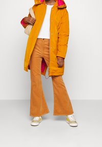 Personne portant un manteau matelassé jaune moutarde avec une doublure rose, un haut blanc, un pantalon pattes d'éléphant en velours côtelé orange, des baskets jaunes, tenant un sac texturé beige.