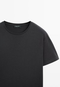 T-shirt noir uni à manches courtes avec col rond, montrant le col et la manche droite sur fond blanc.