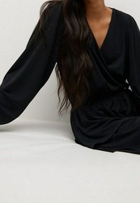 Vestido negro envolvente con mangas largas, tejido suave, escote en V y detalle de cintura fruncida. Modelo sentado sobre un fondo claro.