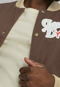 Brown varsity jacket with beige ribbed collar, gold snap buttons, and white embroidered text. Layered over a cream T-shirt.