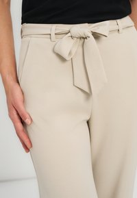 Pantalon beige taille haute avec une ceinture en tissu nouée en nœud à la taille, porté avec un haut noir, et une main posée sur le côté.
