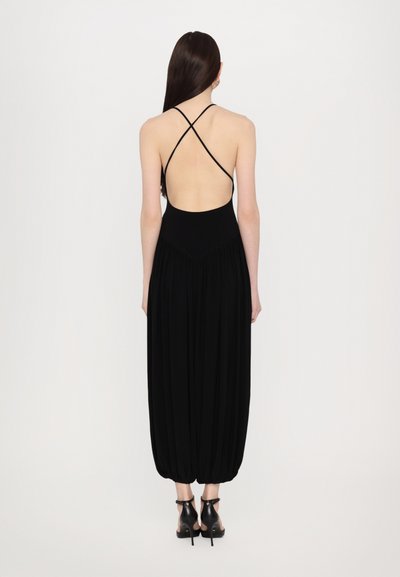 Schwarzer Jumpsuit mit tiefem Rückenausschnitt und überkreuzten Trägern, der ein figurnahes Oberteil und einen lockeren, gerafften Unterteil hat. Glatter Stoff mit matter Oberfläche.