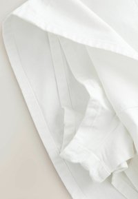 Tissu blanc plié avec des coutures visibles et un bord retroussé, montrant la texture et les détails des points sur un fond clair.