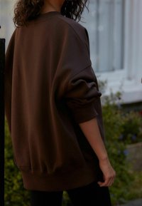 Brown oversized sweatshirt with dropped shoulders, ribbed cuffs, and a soft texture, worn with black leggings.