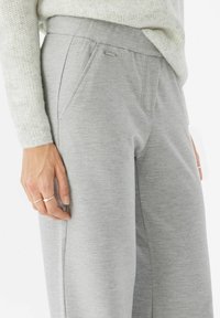 Pantalon de survêtement gris en tissu doux et texturé, avec une ceinture élastique et des poches latérales. Design minimaliste avec un petit accent métallique à l'arrière.