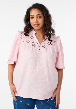 Femme aux cheveux bouclés et lâchés, portant un chemisier rose clair avec des découpes florales et des manches à volants, associé à un pantalon bleu avec de petits nœuds roses.