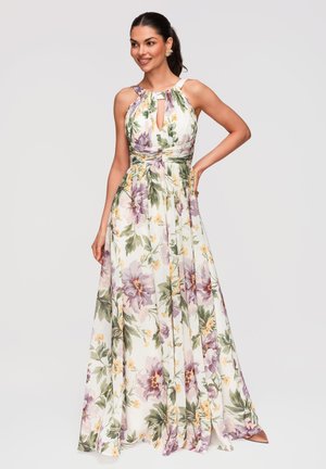 Kvinde iført en hvid, ærmeløs maxikjole med lilla og gule blomster, poserer med den ene hånd på hoften.