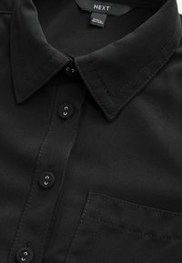Schwarzes Polohemd mit Knopfleiste, ausgestattet mit einer Brusttasche und tonal abgesteppten Nähten. Hergestellt aus glattem Stoff, das Etikett trägt die Aufschrift "NEXT".