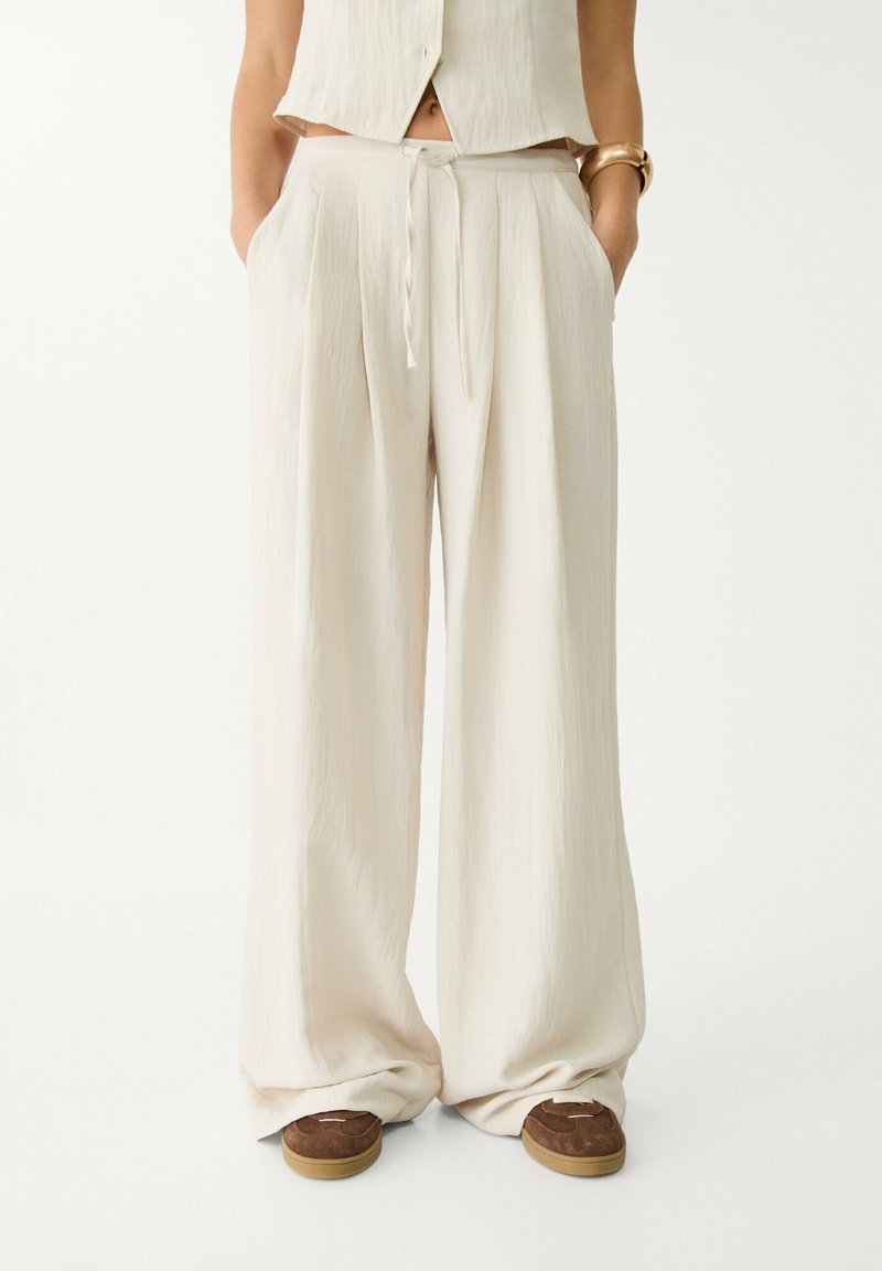 Personne portant un pantalon crème taille haute à jambes larges avec un lien à l'avant et des poches, associé à des chaussures plates marron et un haut crème sans manches.