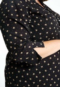 Blouse noire à manches longues avec un petit motif de tête de tigre et des points orange, manches retroussées, portée par une personne aux bras croisés.