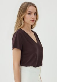 T-shirt a collo V scuro in marrone con maniche corte, realizzato in un tessuto morbido. La maglietta ha una vestibilità rilassata e una consistenza liscia.