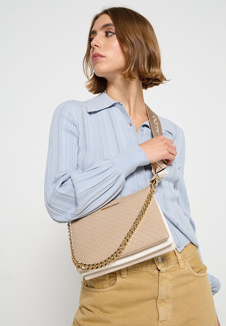 Beige gesteppte Handtasche mit einer goldenen Kettenriemen, die ein strukturiertes Design und glatte Akzente aufweist, getragen über einem hellblauen strukturierten Pullover.