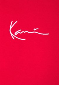 Roter Baumwollstoff mit einem bestickten weißen Signaturdesign. Die Textur ist glatt mit einem gleichmäßigen gerippten Muster.