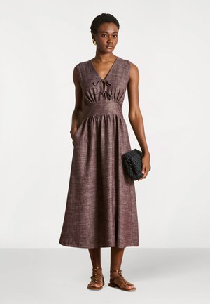 Femme portant une robe midi marron sans manches avec un col noué, tenant une pochette noire texturée et portant des sandales marron à brides, fond neutre.