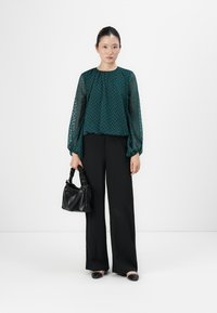 Woman standing, wearing a sheer dark green patterned blouse, black wide-leg pants, black flats, and holding a small black handbag.