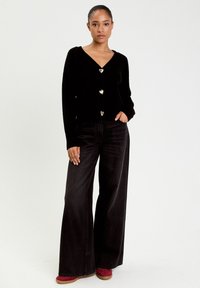 Schwarzer Cardigan mit V-Ausschnitt, drei herzförmigen goldenen Knöpfen, kombiniert mit weit geschnittenen schwarzen Jeans und roten Schuhen mit strukturierten Sohlen.