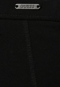 Tessuto nero con una texture liscia presenta un'etichetta metallica con il logo "GUESS" vicino alla parte superiore, evidenziando un design semplice ed elegante.