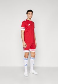 Röd sporttröja och shorts med vita ränder, med en adidas-logotyp. Vita fotbollssockor med blå randiga detaljer och vita fotbollsskor.