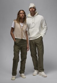 Strikket beige vest med brun kant, hvid t-shirt, oliven cargo bukser og hvid hoodie; mandlig model bærer en hvid beanie. Glat tekstur, afslappet pasform.