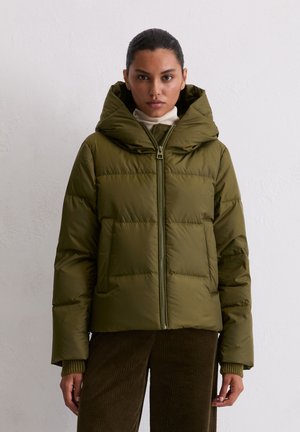 Donna che indossa un giubbotto imbottito verde oliva con colletto alto, un dolcevita bianco e pantaloni in costina marrone, in piedi di fronte a un muro bianco semplice.