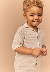 Beige kortærmet strikket cardigan med knapper foran, der har et tekstureret mønster og en krave. Blødt, let materiale.