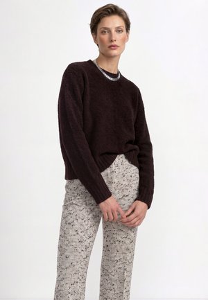 Donna che indossa un maglione marrone scuro con texture e pantaloni chiari con motivi, in piedi davanti a uno sfondo bianco uniforme.