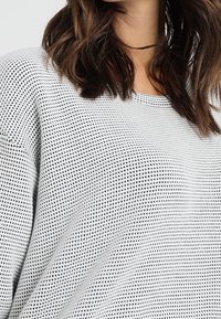 Suéter de punto blanco ligero con un patrón texturizado que presenta pequeños agujeros. Cuello redondo y corte relajado, adecuado para superponer.
