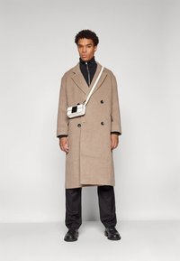 Dvigubai sagstomas rudas vilnonis paltas su dideliais mygtukais, juodas golfas, juodos kelnės ir maža balta per petį nešiojama rankinė su juodu akcentu.