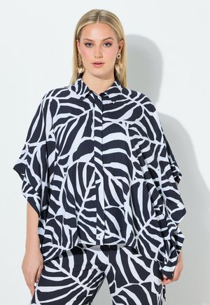 Mujer rubia con camisa suelta y pantalones a juego de patrón abstracto en blanco y negro, con grandes pendientes dorados, sobre fondo blanco.