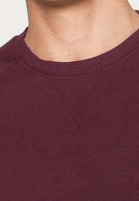 Nærbillede af en nakke og øvre bryst, der bærer en vinrød crewneck-sweatshirt med synlig syningsdetalje under kraven.