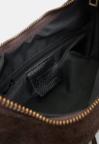 Bruine suede handtas met een interieur van zwarte stof, zichtbare ritszak en een gestructureerd zwart leren label. Gouden ritsdetail aan de rand.