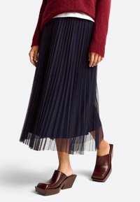 Navy pleated skirt made from lightweight material, layered with a sheer undulating hem. Brown block-heeled mules complement the outfit.