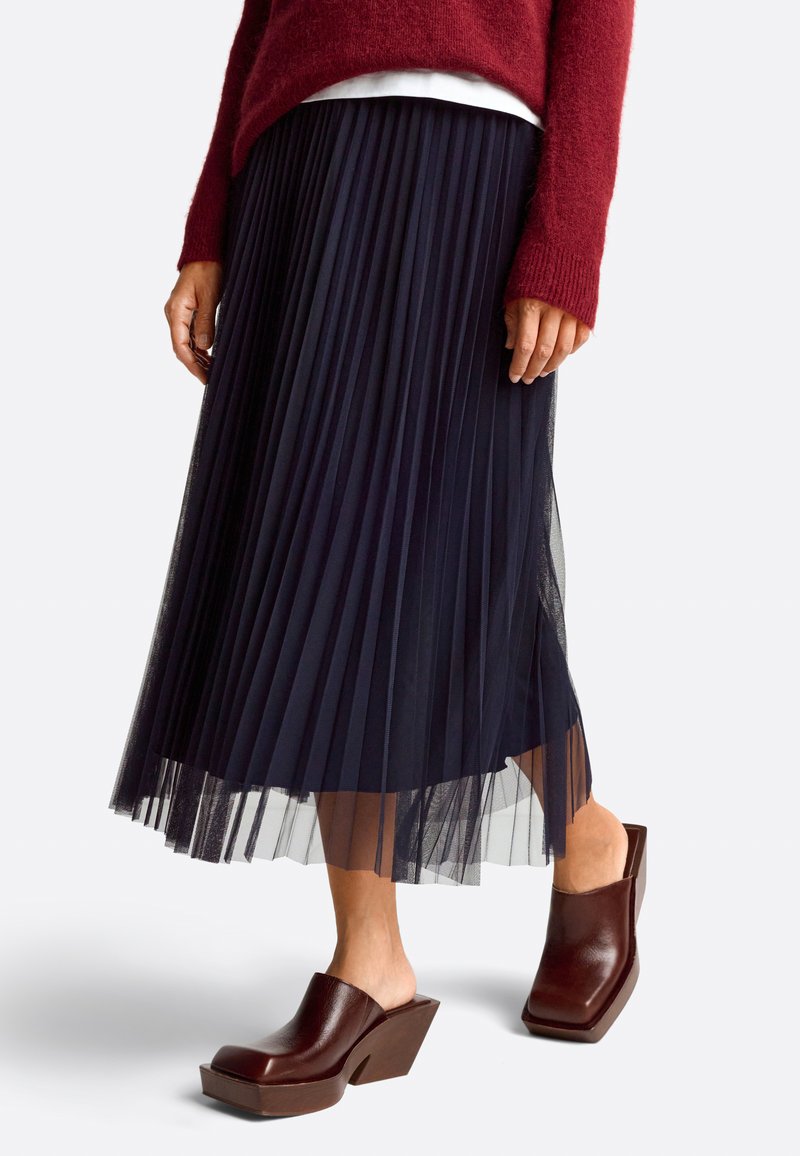 Navy pleated skirt made from lightweight material, layered with a sheer undulating hem. Brown block-heeled mules complement the outfit.