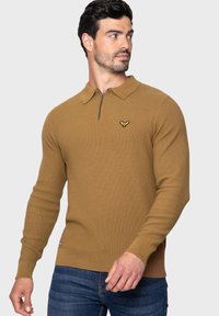Bruine gebreide pullover met een ritskraag, voorzien van een klein geel logo op de borst. Geribbelde manchetten en zoom, textuurpatroon door het hele kledingstuk.