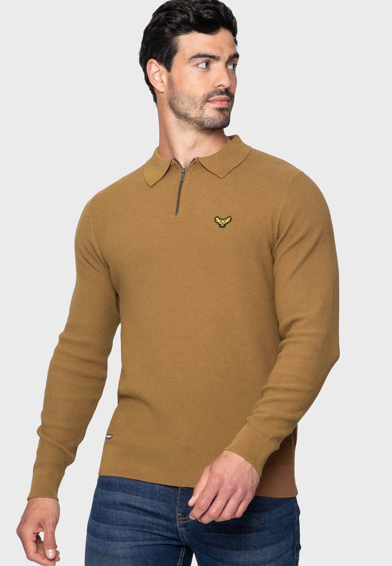 Bruine gebreide pullover met een ritskraag, voorzien van een klein geel logo op de borst. Geribbelde manchetten en zoom, textuurpatroon door het hele kledingstuk.