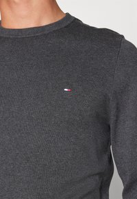 Närbild av en person som bär en mörkgrå stickad tröja med en liten rektangulär röd, vit och blå logotyp på bröstet.