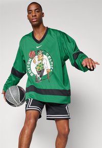 Zöld, hosszú ujjú mez, amelyen Boston Celtics grafika található, fekete részletekkel és V-kivágással. Fekete sorttal és egy kosárlabdával párosítva.