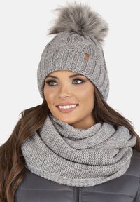 Berretto in maglia grigio con pompon in pelliccia sintetica e design texturizzato, abbinato a una sciarpa a cerchio in maglia grigia coordinata.